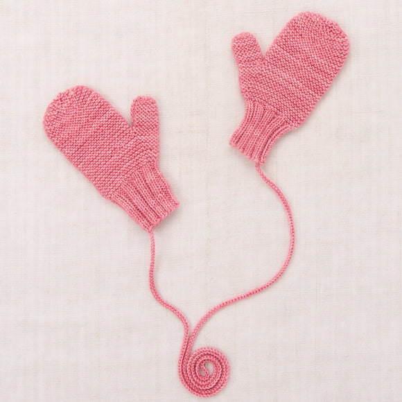 A pair of pink hand-knit baby mittens with a long yarn tail coiled at the bottom, laid flat on a textured off-white linen background.