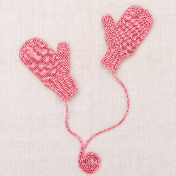 A pair of pink hand-knit baby mittens with a long yarn tail coiled at the bottom, laid flat on a textured off-white linen background.