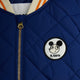 Close-up of a royal blue quilted jacket with a gold zipper, featuring a circular white patch with a black cartoon face and the text 'M. RODINI' on the chest.
