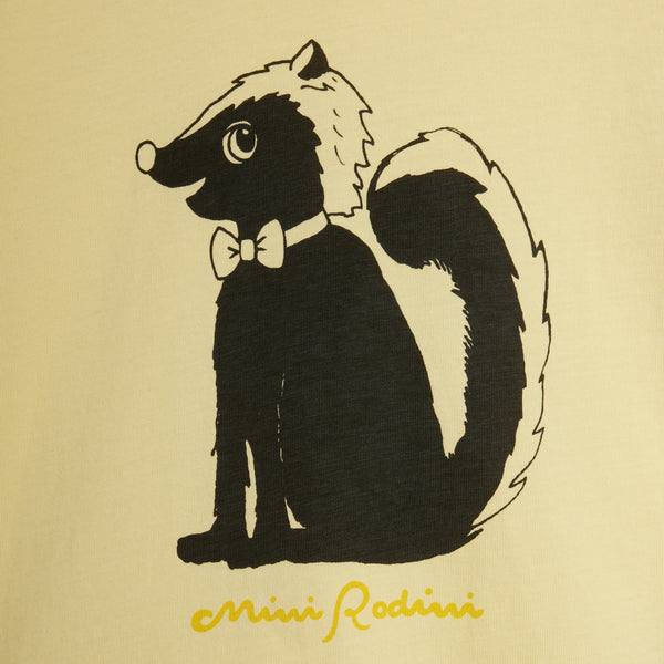 A close-up of a cream-colored fabric surface featuring a black graphic illustration of a badger wearing a white bow tie, with yellow cursive text below reading 'Mini Rodini'.