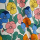 A close-up of a fabric with a colorful pansy floral print on a white background, featuring blue, pink, yellow, and orange flowers with green leaves.