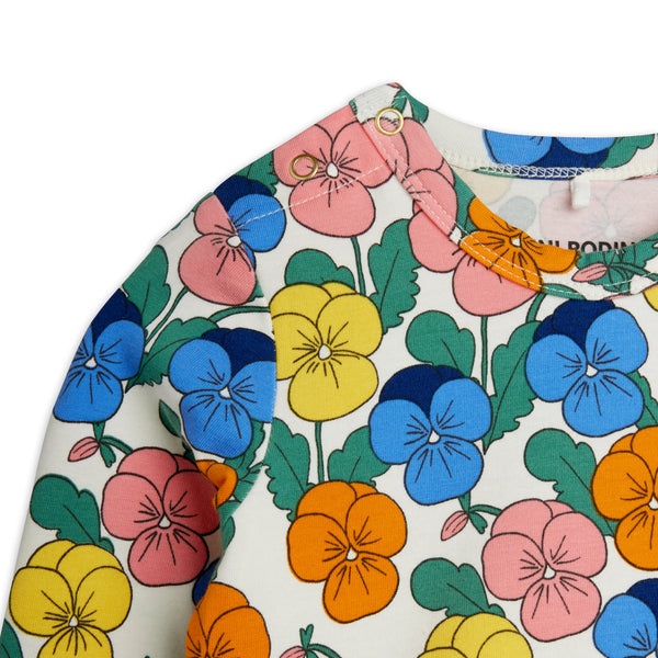 A close-up of a children's long-sleeve top featuring a colorful pansy floral print on an off-white background. The print includes pink, blue, yellow, and orange flowers with green leaves.