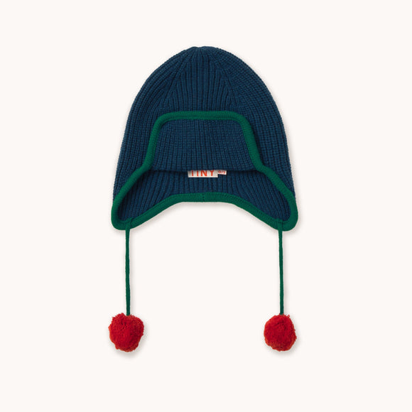 A dark navy blue ribbed knit beanie hat with a green trim around the ear flaps and two red pom-poms hanging from the ear ties, shown against a plain off-white background.