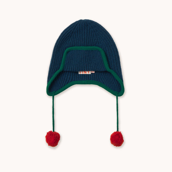 A dark navy blue ribbed knit beanie hat with a green trim around the ear flaps and two red pom-poms hanging from the ear ties, shown against a plain off-white background.