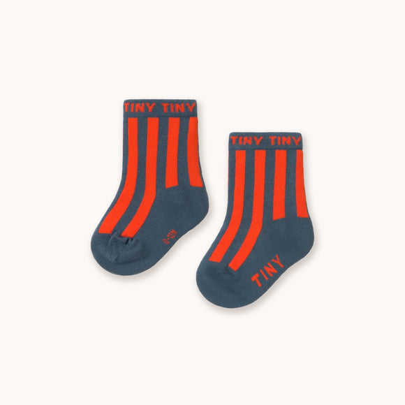 A pair of navy blue baby socks with bright orange vertical stripes and 'TINY' printed in orange on the cuff and sole, displayed on a clean off-white background.