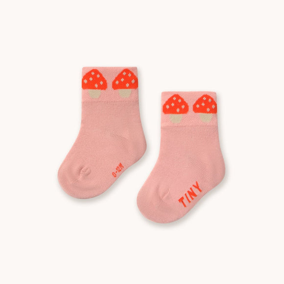 A pair of baby socks in a soft pink color, featuring a red strawberry graphic at the top cuff and the word 'TINY' printed in red on the sole, set against a plain off-white background.