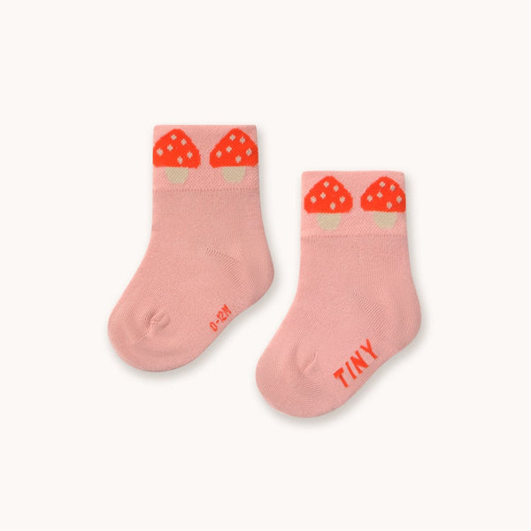 A pair of baby socks in a soft pink color, featuring a red strawberry graphic at the top cuff and the word 'TINY' printed in red on the sole, set against a plain off-white background.