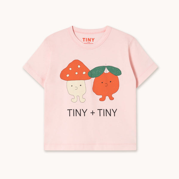 A light pink short-sleeve kids' t-shirt featuring a cute graphic of two tiny cartoon characters, one with a red mushroom cap and one with an orange body and green leaf cap, with the text 'TINY + TINY' printed below. The brand name 'TINY' is visible on the inner neck label.