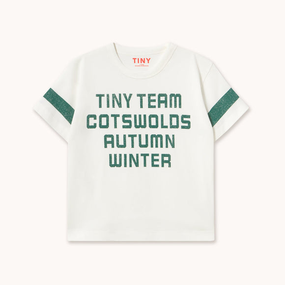 A white short-sleeve T-shirt with green text reading 'TINY TEAM COTSWOLDS AUTUMN WINTER' on the front and green sleeve stripes, shown on a light beige background.