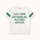 A white short-sleeve T-shirt with green text reading 'TINY TEAM COTSWOLDS AUTUMN WINTER' on the front and green sleeve stripes, shown on a light beige background.