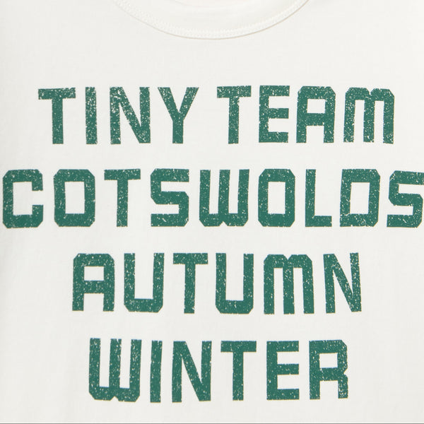 Close-up of a white T-shirt with large distressed green text reading 'TINY TEAM COTSWOLDS AUTUMN WINTER' and a small red 'TINY' neck print.