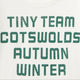 Close-up of a white T-shirt with large distressed green text reading 'TINY TEAM COTSWOLDS AUTUMN WINTER' and a small red 'TINY' neck print.