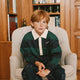 A young boy with red hair sits in a beige armchair in front of a wooden bookshelf. He is wearing a green and navy striped long-sleeve rugby shirt with a white collar, dark blue jeans, and striped socks.