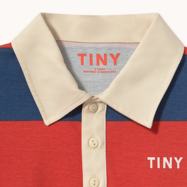 Close-up of a striped polo shirt collar and button placket with navy, red, and cream horizontal stripes. The inside collar shows the brand name 'TINY' in red, and 'TINY' is printed in white on the chest.