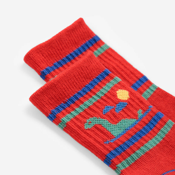 A close-up of a red knit sock with blue and green horizontal stripes and a small dinosaur graphic, photographed on a clean white background.
