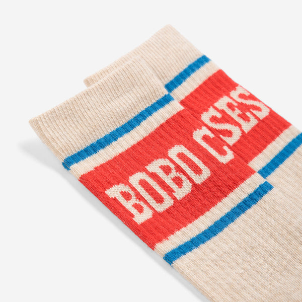 A close-up of a ribbed knit sock in cream with a bold red band, blue stripes, and white 'BOBO CSES' lettering, photographed on a light gray background.