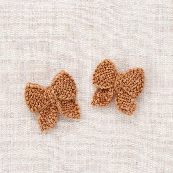 A pair of small crocheted butterfly-shaped hair clips in a warm camel brown color, displayed on a textured off-white linen background.