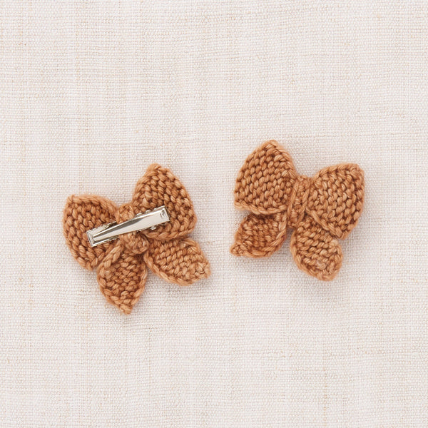 Two small crocheted bow hair clips in a warm tan-brown color, displayed on a textured off-white linen background.