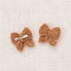 Two small crocheted bow hair clips in a warm tan-brown color, displayed on a textured off-white linen background.