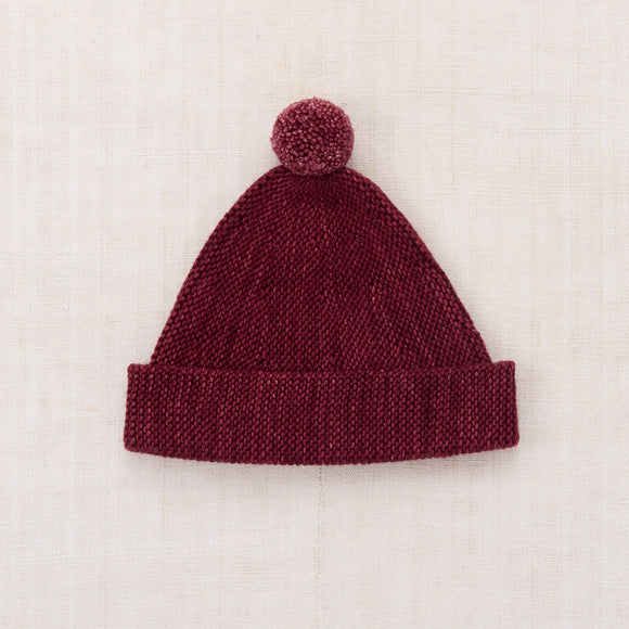A burgundy knit beanie hat with a matching pom-pom, laid flat on a textured off-white fabric background.