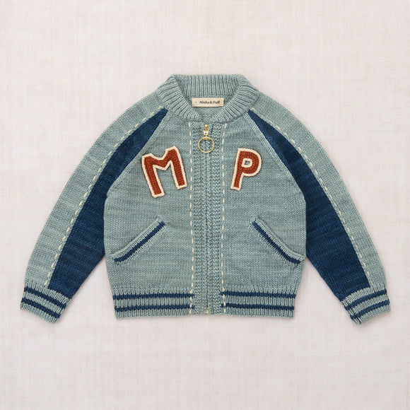 A light blue knit zip-up cardigan with navy blue raglan sleeves and ribbed trim, featuring large red embroidered letters 'M' and 'P' on the chest, laid flat on a light textured background.