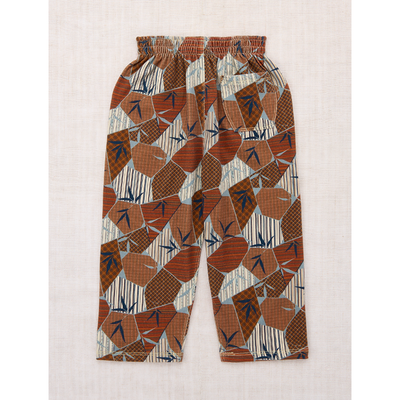 A pair of children's elastic-waist pants with an all-over geometric print in rust orange, brown, cream, and navy blue, laid flat on a light beige textured fabric background.