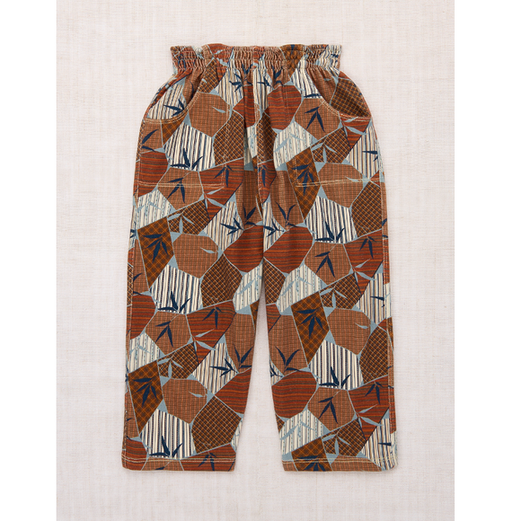 A pair of children's elastic-waist pants with an abstract geometric print in terracotta, brown, beige, and navy blue, laid flat on a textured beige fabric background.