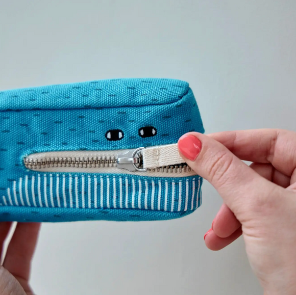 A light blue fabric zipper pouch with a cute cartoon face and a silver zipper being pulled open by a hand with pink nail polish, against a plain light gray background.