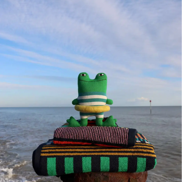 A green plush frog toy wearing a striped green and white shirt and yellow ring float sits on top of folded striped towels at the edge of a beach, with the ocean and blue sky in the background.
