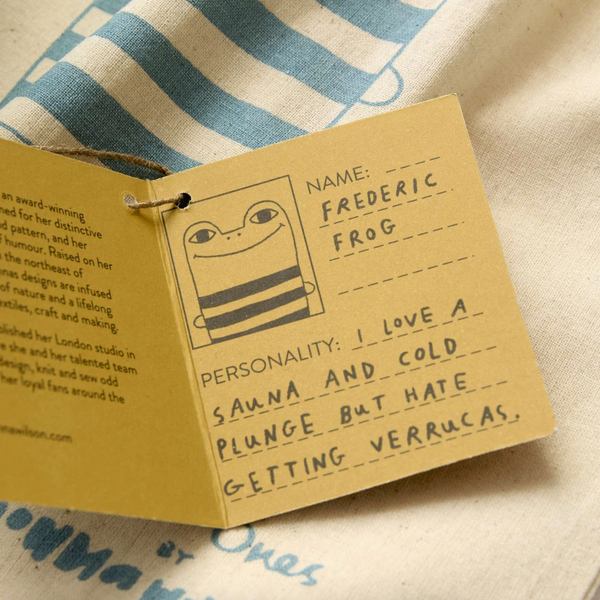 A small kraft paper tag attached to a string, printed with a cartoon frog and handwritten text including the name 'FREDERIC FROG' and a personality statement, laid on a cream fabric with blue printed text.