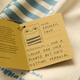 A small kraft paper tag attached to a string, printed with a cartoon frog and handwritten text including the name 'FREDERIC FROG' and a personality statement, laid on a cream fabric with blue printed text.
