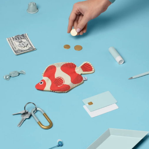 A fish-shaped fabric pouch with red and white vertical stripes lies on a light blue surface, surrounded by styled props including keys, a carabiner, cards, a pen, coins, and a hand placing a small white object near the top.