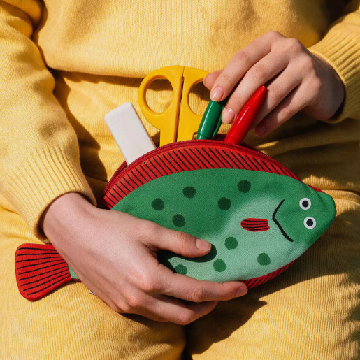 A person in a yellow sweater holds a fish-shaped green and red pencil pouch with black polka dots, holding yellow, green, and red pens inside.