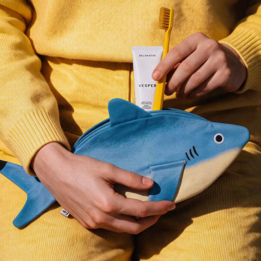 A person in a yellow sweater holds a blue and cream shark-shaped zip pouch while inserting a yellow toothbrush and a white Vesper tube into the pouch.