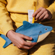 A person in a yellow sweater holds a blue and cream shark-shaped zip pouch while inserting a yellow toothbrush and a white Vesper tube into the pouch.