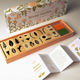 A boxed stamp garden set displayed on a light neutral background, with the floral-patterned box behind an open wooden tray holding multiple wooden rubber stamps and two small color blocks. An open booklet with printed instructions lies in front.