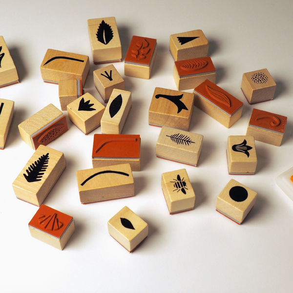 A set of small wooden rubber stamp blocks scattered on a white surface, each featuring different engraved or stamped botanical and abstract designs in black and reddish-brown.