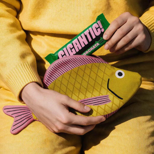 A person in a yellow knit outfit holds a fish-shaped coin purse in their hands while holding a green Filaicant! chocolate bar above it. The coin purse is olive green with pink stripes and a pink top opening.