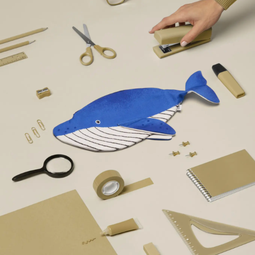A whale-shaped desk organizer made from blue and white striped cardstock lies on a beige tabletop surrounded by stationery items, including a hand using a stapler, scissors, tape, a magnifying glass, and rulers.
