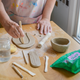 A person wearing a paint-splattered apron works at a wooden table with clay tools, a rolling pin, a small clay bowl, and a green bag of clay. Their hands are shown shaping a piece of clay.