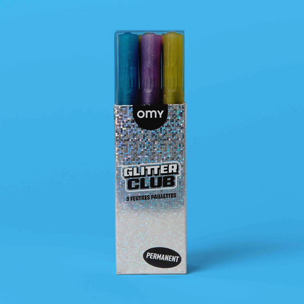 A boxed set of three Omy Glitter Club permanent makeup pens on a solid blue background. The pens are visible through a clear window at the top of the silver glitter box.