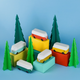 A set of colorful wooden toy camper vans arranged on pastel blocks with green tree shapes against a light blue background.